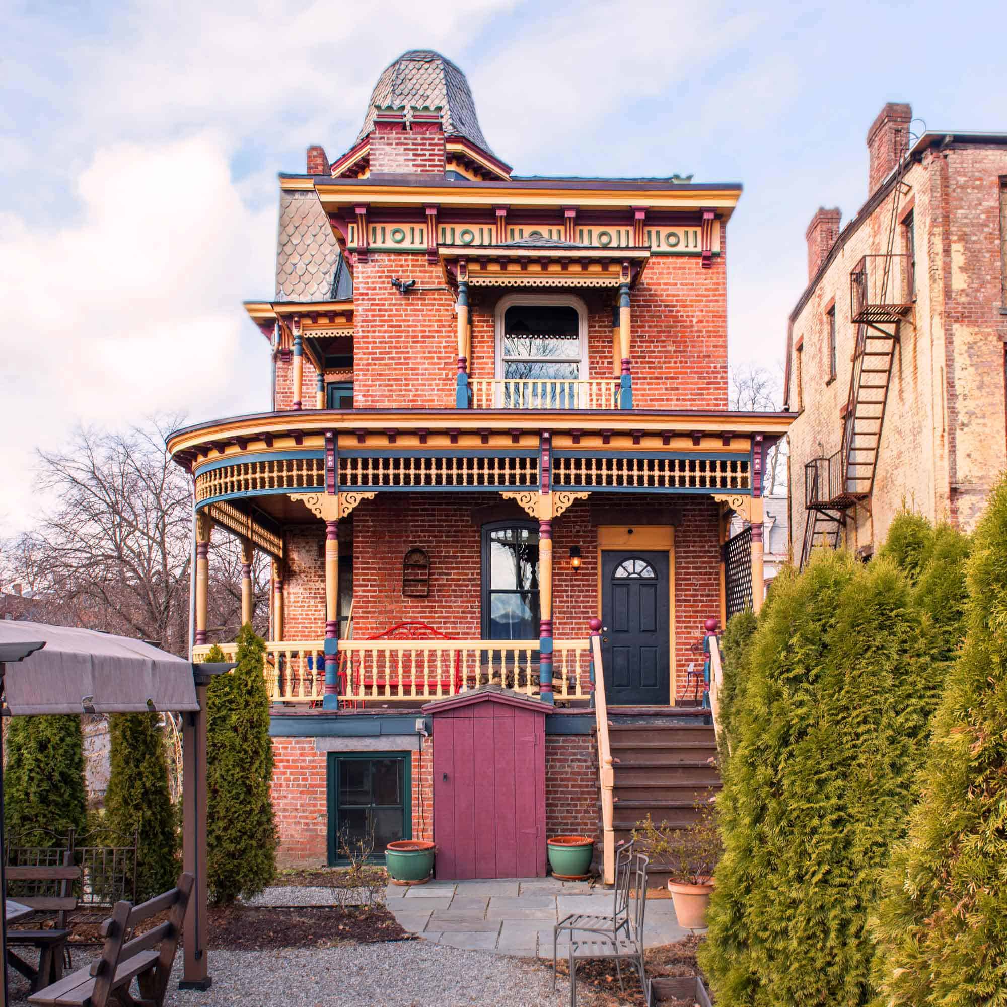 The rest view of a Brick Victorian with elaborate porches, roof brackets, and a mansard roof. The wooden details are rendered in shades of blue, yellow, and maroon.