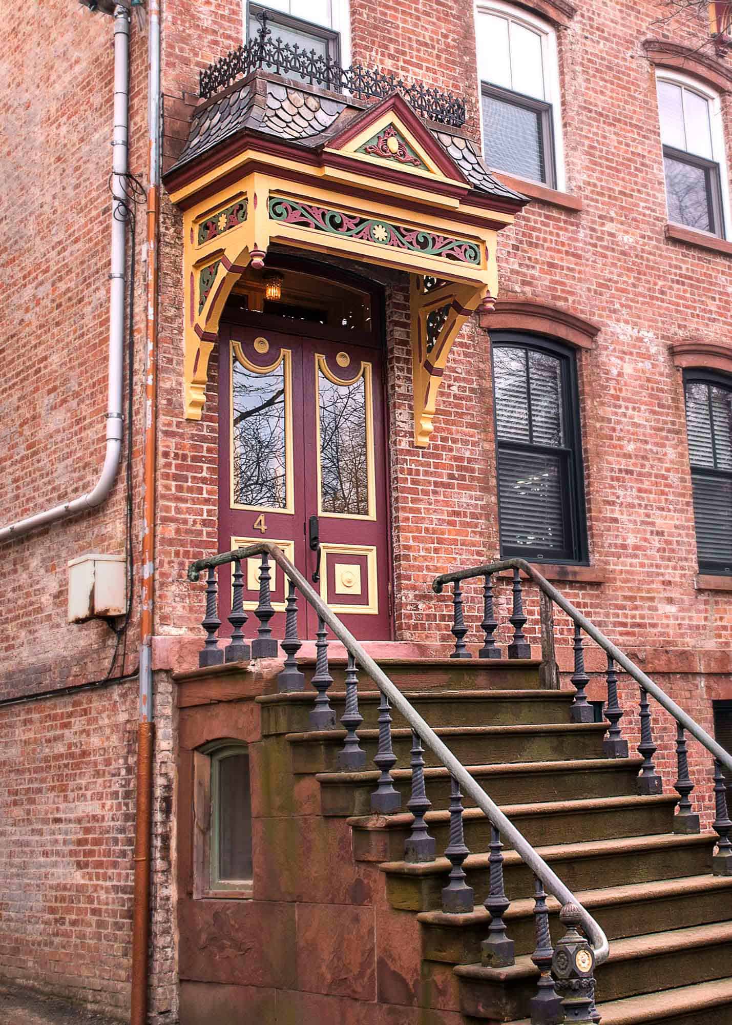 The front stoop of a Second Empire Brick Building in Newburgh, New York. Steps lead up to he front entrance, with iron railings on either side. Above is a highly-detailed wooden overhang with slate tile miniature roof, and a Classic Newburgh double door.