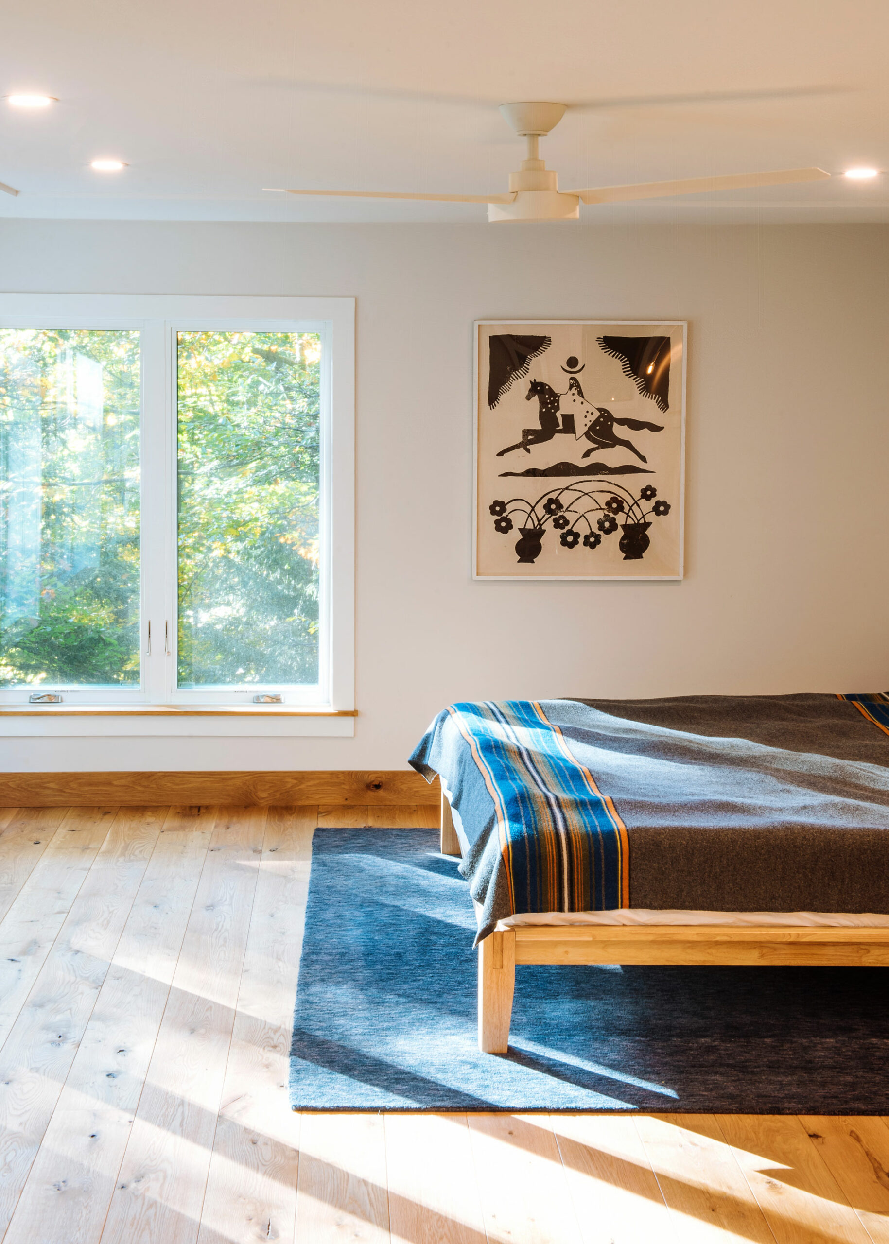 Master bedroom with slanting light coming in through windows off the edge of the frame. To the right is a low platform bed and a grey carpet. Behind is a framed black and white print.