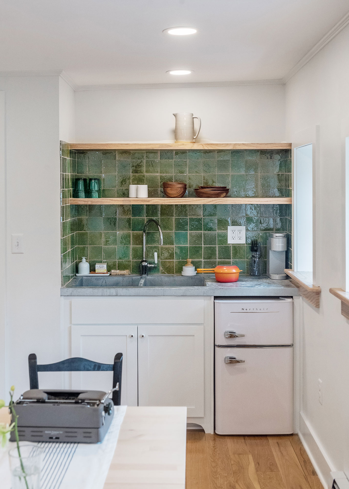 A green-tiled kitchenette with a small retro-style under-counter fridge, custom shelves, and a green tile backsplash.